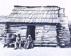 an old photo of two men sitting in front of a log cabin with a thatched roof