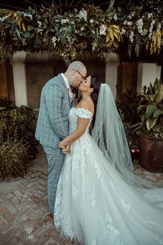 a bride and groom kissing in front of an arch with greenery on the wall