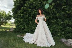 a woman standing in front of a bush wearing a wedding dress with off the shoulder sleeves