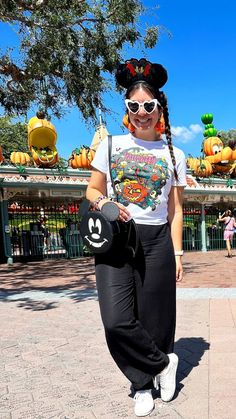 Julia Vaness wearing black linen pants with a white graphic tshirt from Disneyland 2023 Halloween collection, with loungefly mickey mouse bat bag, pumpkin plush earrings, white heart shaped sunglasses and black fuzzy halloween mickey ears