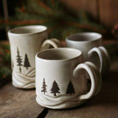 two coffee mugs sitting on top of a wooden table next to pine tree branches