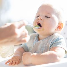 a baby sitting in a highchair eating food from a spoon while being held by someone's hand