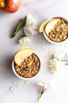 two white bowls filled with granola next to an apple and flowers on a table