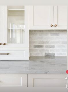 a kitchen with white cabinets and marble counter tops in front of a gray brick wall