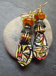 two pairs of earrings sitting on top of a rock next to each other, one with an orange and yellow glass bead hanging from it