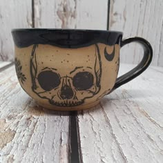 a coffee cup with a skull painted on the front and side is sitting on a wooden table