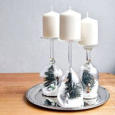 three white candles are sitting on a silver plate with snow - covered pine trees in them