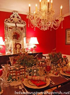 a dining room table set for christmas with red walls and chandelier in the background