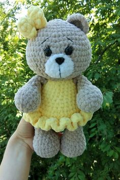 a crocheted teddy bear in a yellow dress is held up by someone's hand