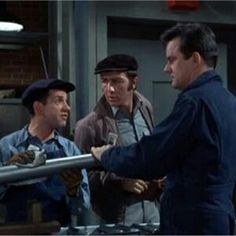 three men are talking to each other in an industrial setting, one is wearing a blue jacket and the other wears a black hat