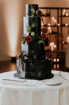 a three tiered black wedding cake with red flowers on the side and candles in the background