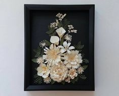 a black frame with white flowers and greenery in it is hanging on the wall