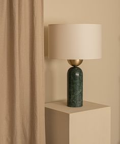 a green vase sitting on top of a white table next to a lamp and curtain