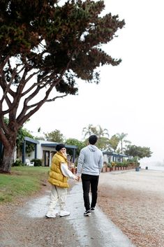 two people holding hands walking down a path in front of a tree on a cloudy day