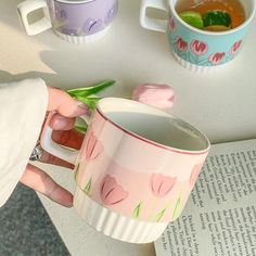 a woman is holding a coffee mug with flowers on it next to two cups and an open book