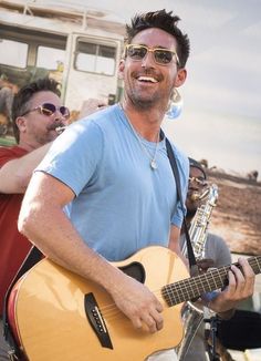 a man holding an acoustic guitar while standing next to another man with sunglasses on his head