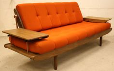 an orange futon sofa with wooden legs and arm rests against a white brick wall