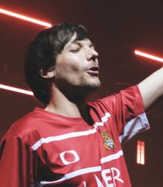 a man in a red shirt is holding his hand out to the side while singing into a microphone