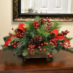 a christmas arrangement with pine cones, holly and red berries on a wooden table in front of a mirror