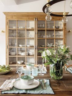 a dining room table set with plates, glasses and flowers in a vase next to the china cabinet