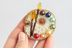 a hand holding a beaded brooch with paintbrushes and beads on it