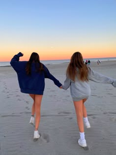 two girls walking on the beach holding hands and looking at the sunset in the distance