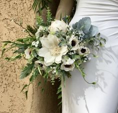 a bride's bouquet with white flowers and greenery on her wedding day in front of a stucco wall