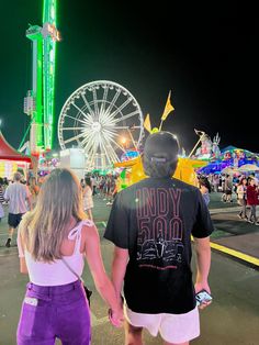 two people holding hands in front of an amusement park ferris wheel and carnival rides at night