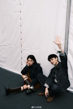 two people sitting on the ground in front of a white wall with their hands up