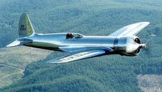 an airplane flying in the sky over some trees and land with mountains in the background