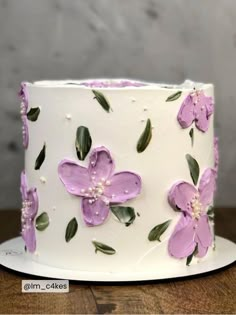 a white cake with purple flowers and green leaves on the top is sitting on a wooden table