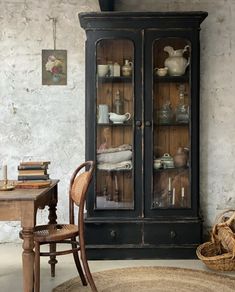 an old fashioned china cabinet with glass doors and shelves in the corner, next to a wicker chair