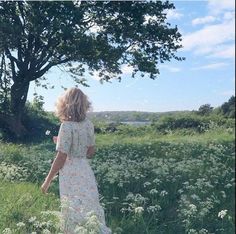 a woman in a floral dress is walking through tall grass near a tree and water