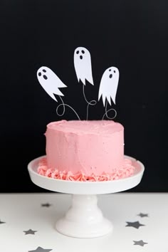 a pink cake topped with three ghost cutouts on top of a white cake plate