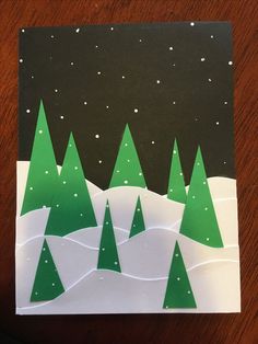 a card with green trees on it and snowing in the sky behind them, sitting on a wooden table