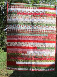 a red and white quilt hanging from a clothes line in front of some trees on a sunny day