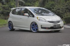 a white car with blue rims drives down the street in front of some trees