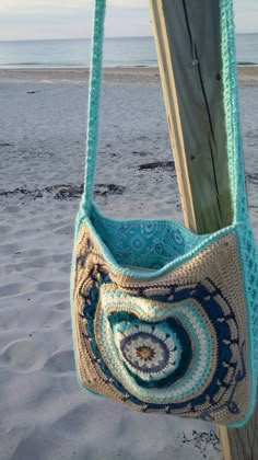 a crocheted bag hanging from a wooden post on the beach with water in the background