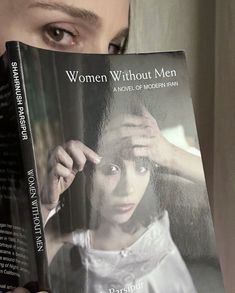 a woman holding up a book about women without men in front of her face and looking at the camera