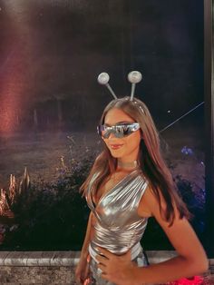 a woman in silver dress standing next to a wall with lights on her head and sunglasses on