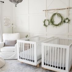 two white cribs sitting in a room next to a chair and wall decoration