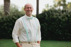 a man wearing a bow tie and suspenders standing in front of some bushes smiling