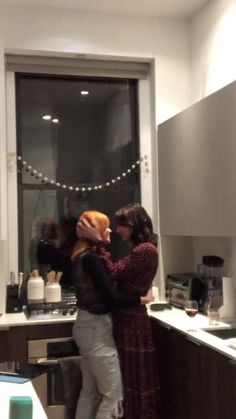 two people standing in a kitchen hugging each other with lights on the wall behind them