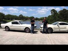 two men standing next to white mustangs in front of a body of water and trees