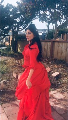 a woman in a long red dress standing on the ground next to some bushes and trees