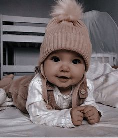 a baby laying on top of a bed wearing a knitted hat and jacket with fur pom poms