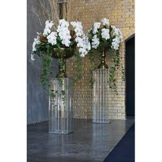 three clear vases with white flowers in them on a gray floor next to a brick wall