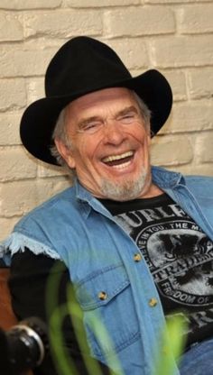 an older man wearing a cowboy hat sitting in front of a brick wall and smiling