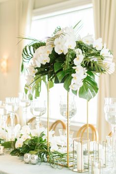 the table is set with white flowers and greenery in tall gold vases, along with candles