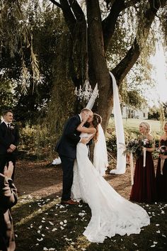 a bride and groom kissing under a tree with their wedding party in the back ground